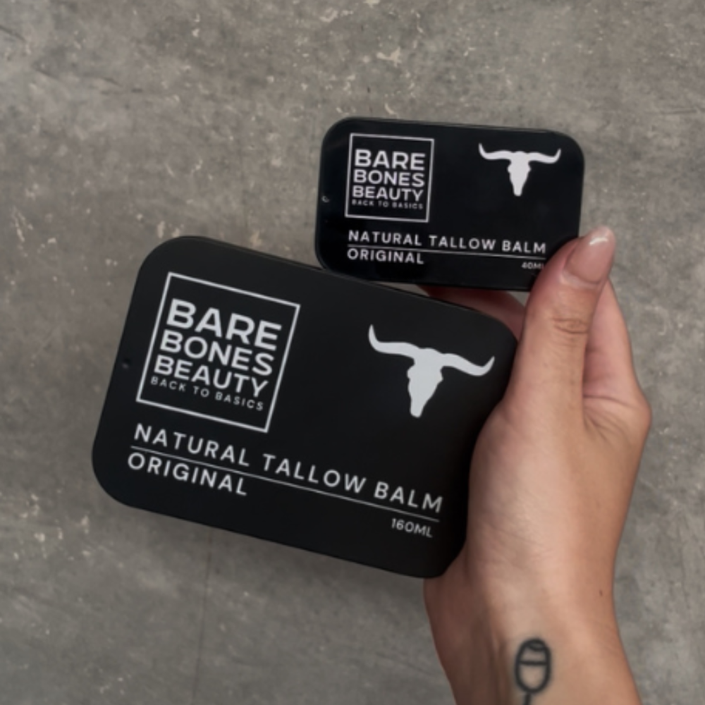 
                  
                    A hand with a small tattoo holds two black tins of Bare Bones Beauty Australia Original Tallow Balm (160mL and 40mL), showcasing skin hydration against a textured gray background.
                  
                