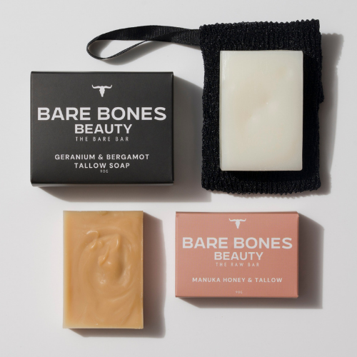 Bare Bones Beauty Australia’s Soap Duo—one white, one beige—lies beside branded boxes. The black box features geranium & bergamot; the pink spotlights Manuka honey and grass-fed beef tallow. The white bar rests on a black pouch.