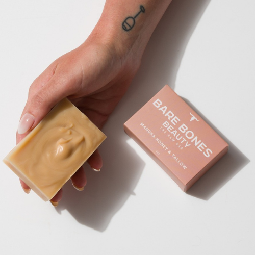 A hand with a small tattoo holds The RAW Bar – Tallow Soap by Bare Bones Beauty Australia, made with grass-fed beef tallow and Manuka Honey, next to its pink box on a white background.