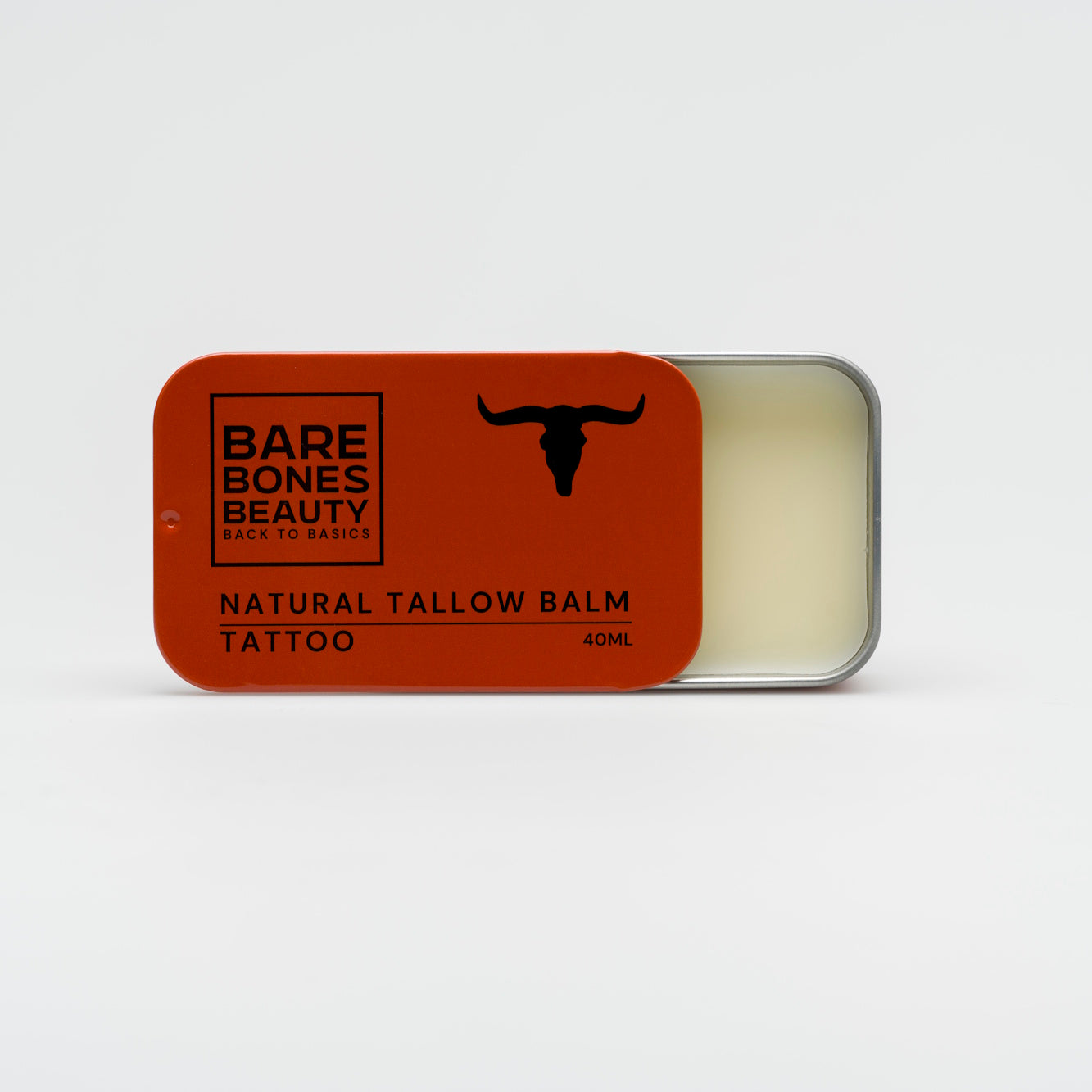 A partially open rectangular tin of Bare Bones Beauty Australia Tattoo Balm shows the balm inside. The orange lid has black text and a bull's head silhouette. Made with natural ingredients to support skin repair.