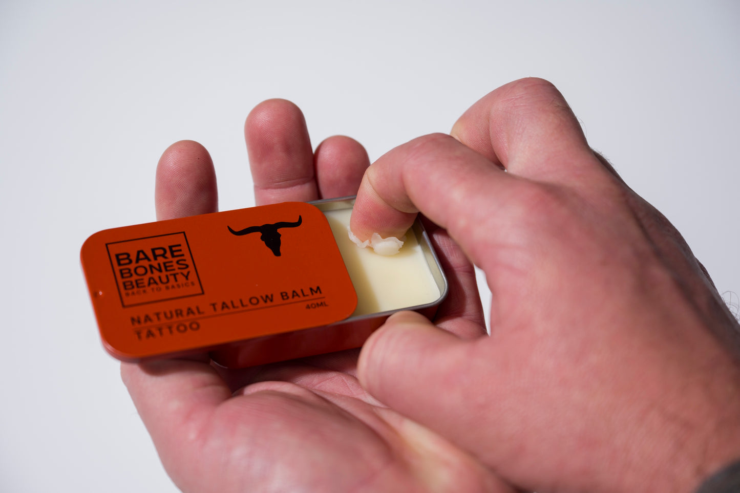 A person scoops Tattoo Balm by Bare Bones Beauty Australia, a natural skin repair balm, from a rectangular tin with an orange bull-logo lid—one hand holding the tin while the other applies the product.