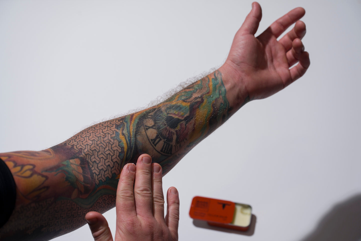 A tattooed arm with colorful designs, including a clock, is shown as another hand applies Bare Bones Beauty Australia Tattoo Balm. The natural balm comes in a small rectangular orange tin resting on the white surface below.