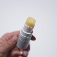 A hand holds a Bare Bones Beauty Australia Tallow Lip Balm (4 for $40) with the cap off, revealing the pale yellow balm. The plain white background highlights this natural essential for lasting lip hydration.